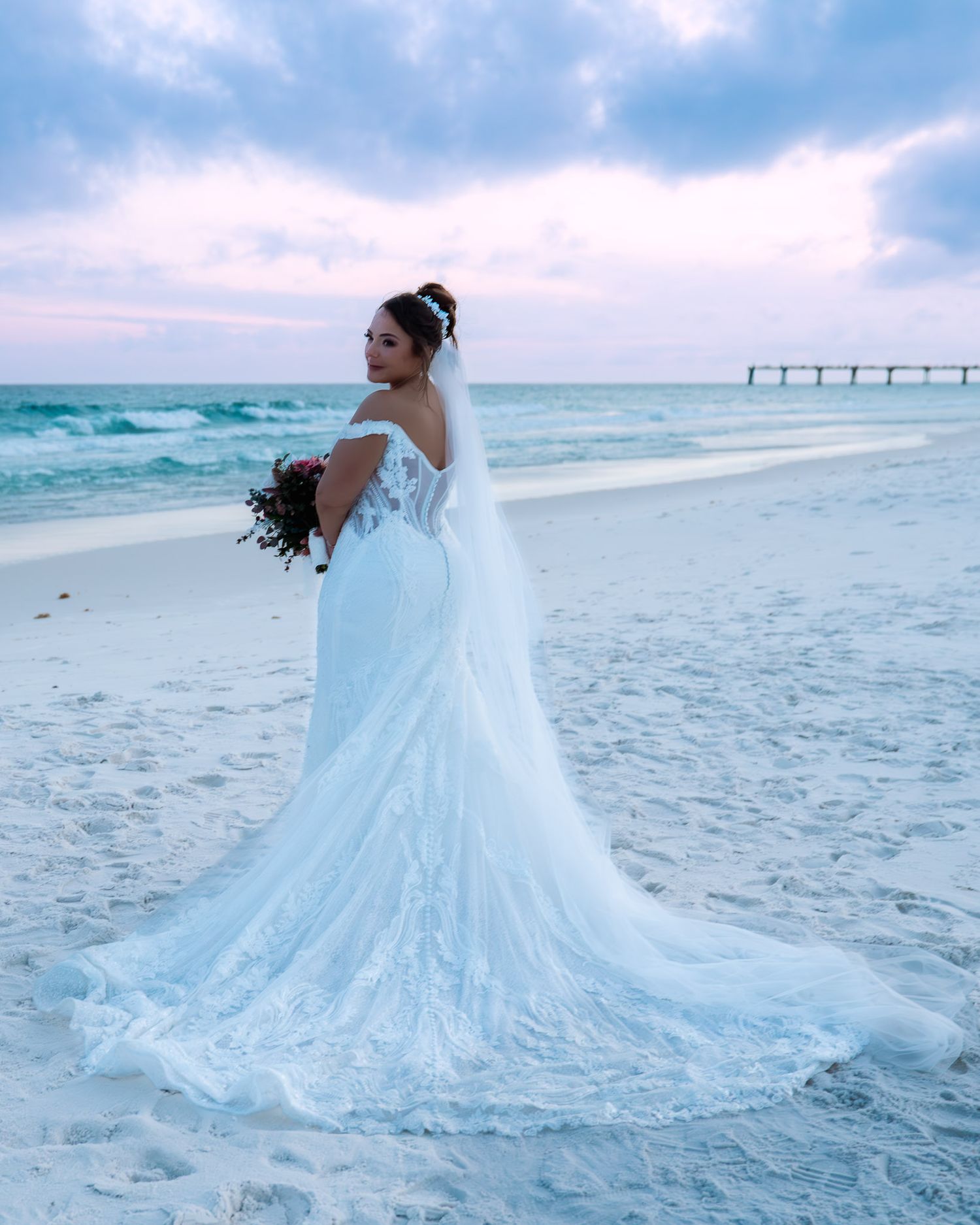 Bride in elegant lace off-shoulder wedding gown with veil stands on a sandy beach at sunset near a pier.