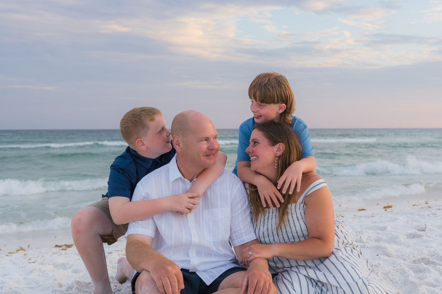 Family of four smiling together on a sandy beach at sunset, with ocean waves in the background.