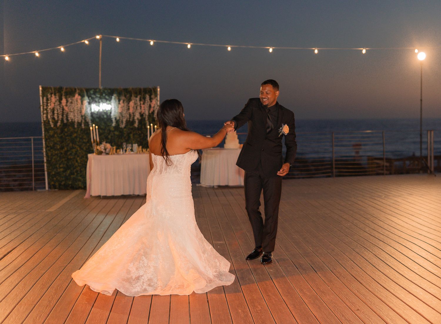 Newlyweds dance together on a waterfront deck at twilight with string lights glowing in the background.