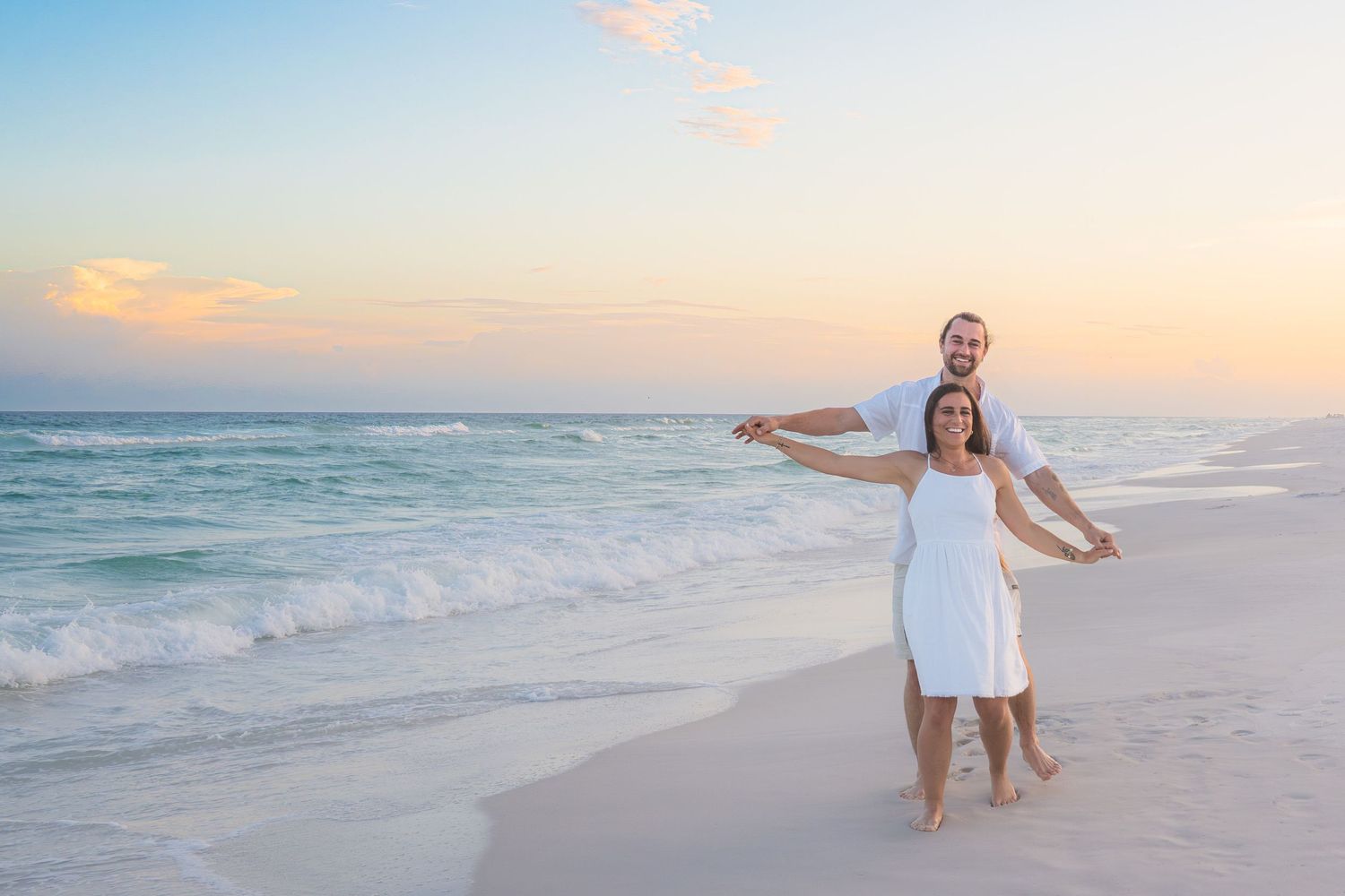 Couple in white outfits dancing on a sandy beach at sunset with turquoise ocean waves in the background.
