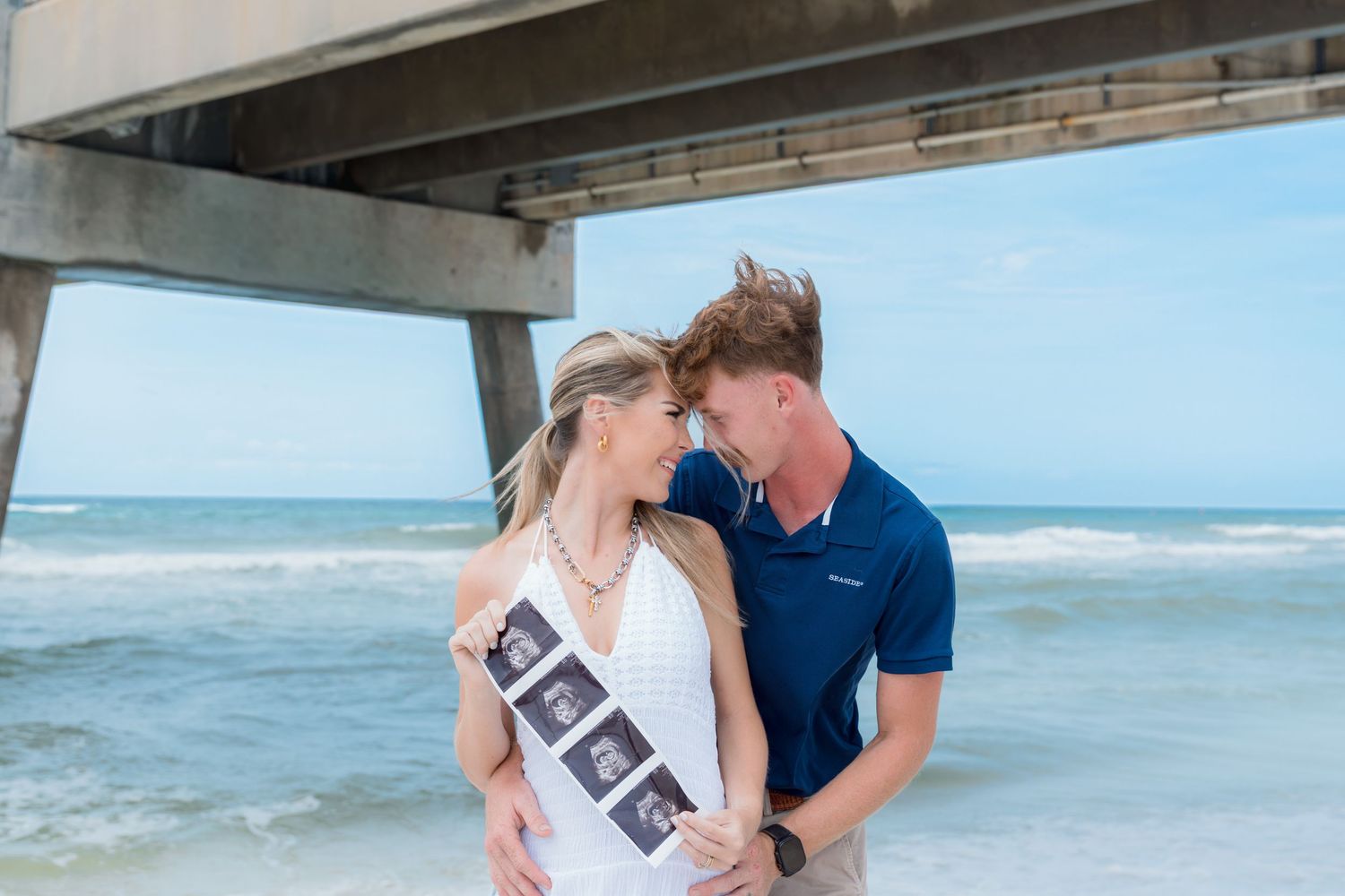 Couple embracing on beach holding ultrasound photos, pregnancy announcement near ocean pier.