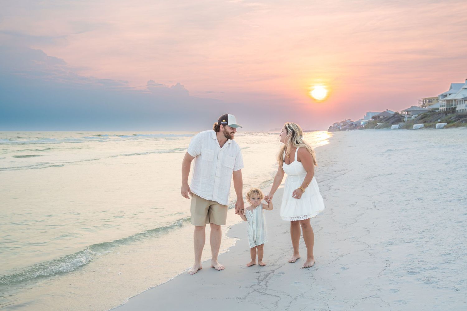 Family of three holding hands on a sandy beach at sunset, wearing coordinated white outfits with pastel sky backdrop.