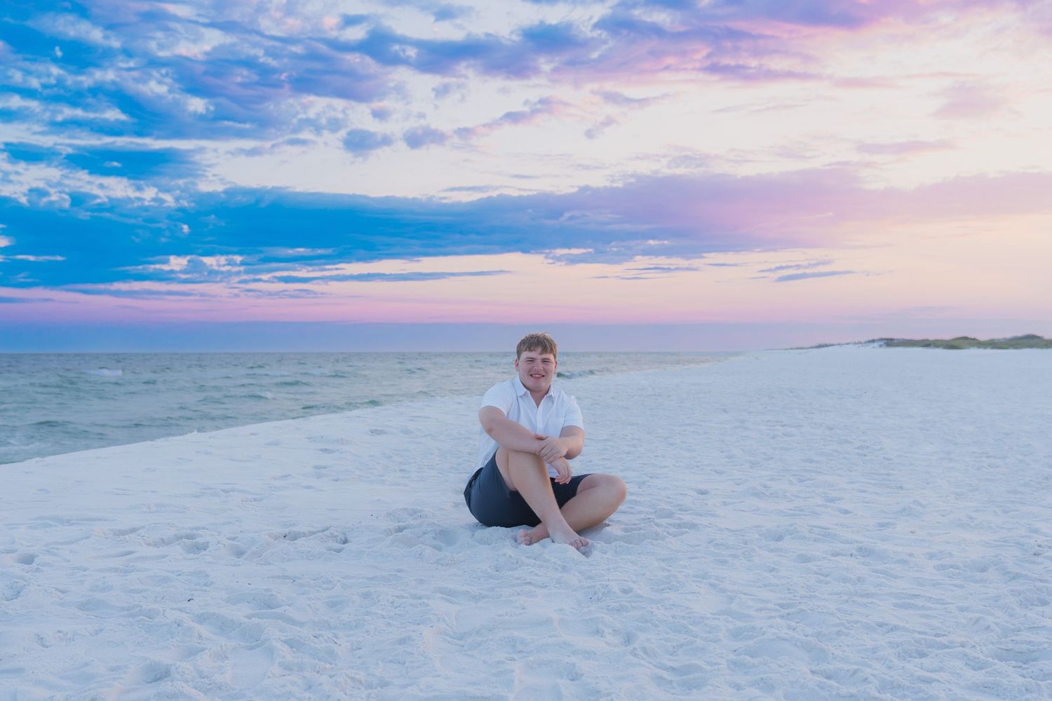 Person sitting cross-legged on white sand beach at sunset with colorful pink and blue sky over calm ocean.