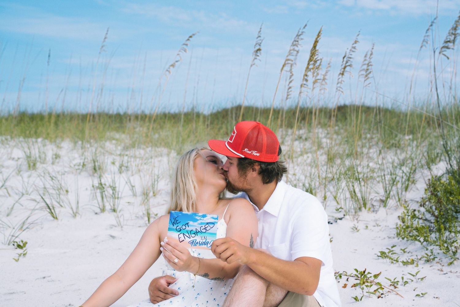 Couple kissing on white sand beach dunes wearing red caps, romantic beach engagement photo with sea oats in background.