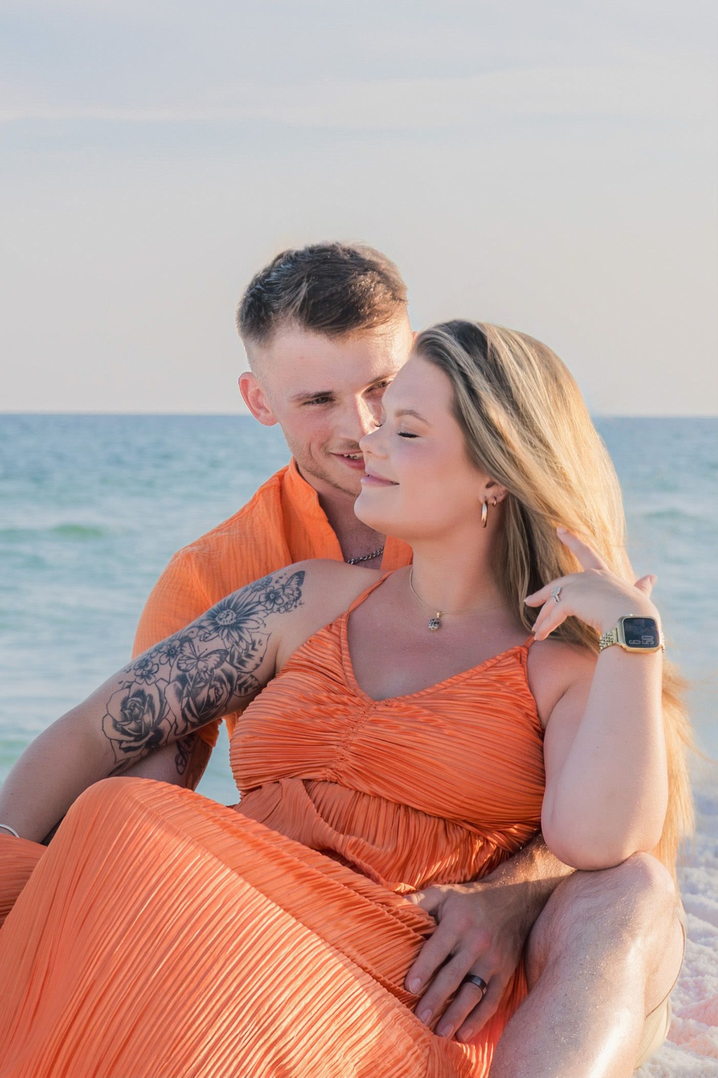 Couple in matching orange outfits sitting together on a beach, man with tattoos embracing woman with long blonde hair.