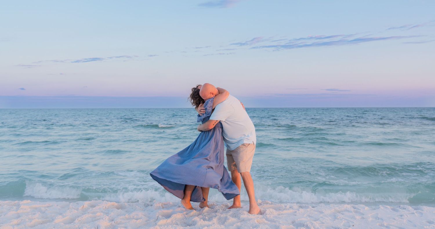 Couple embracing romantically on a sandy beach at sunset with gentle ocean waves and pastel sky.