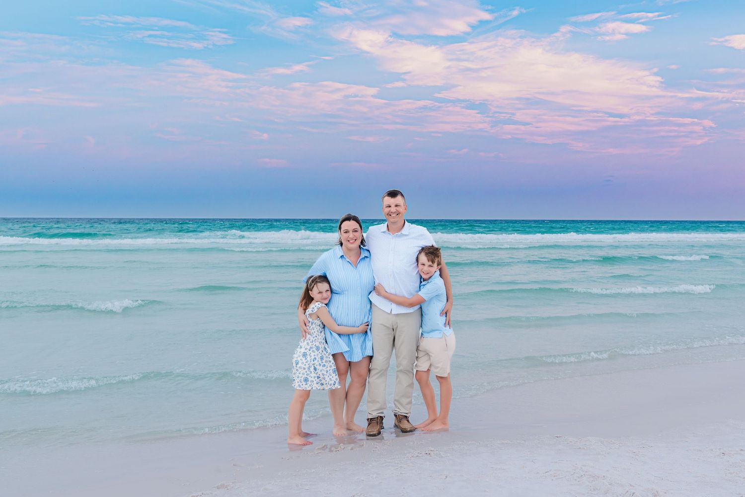 Family of four poses on a turquoise beach at sunset with pastel pink and blue sky backdrop.