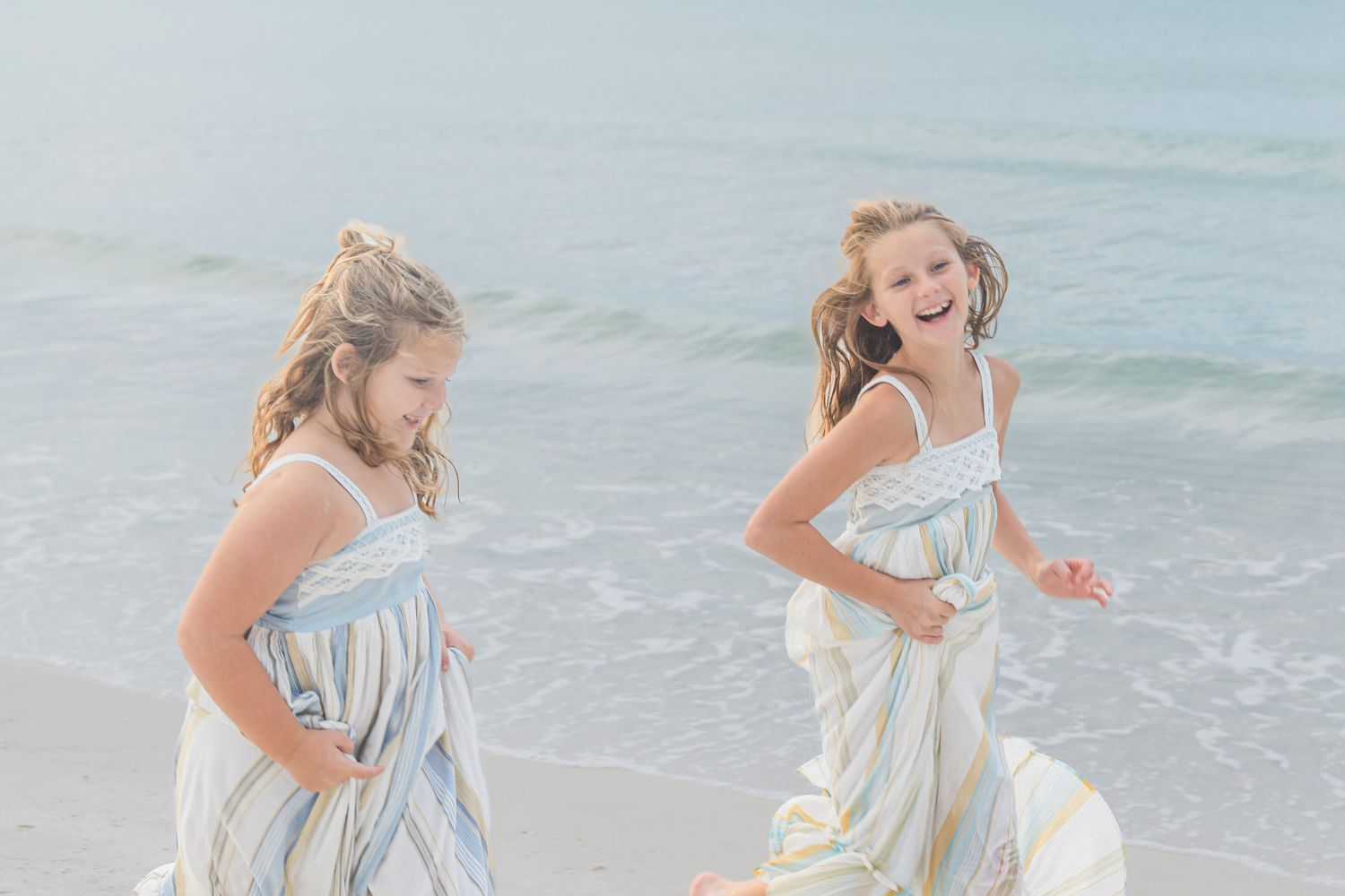 Two girls in striped maxi dresses laugh and play on a sunny beach with gentle ocean waves behind them.