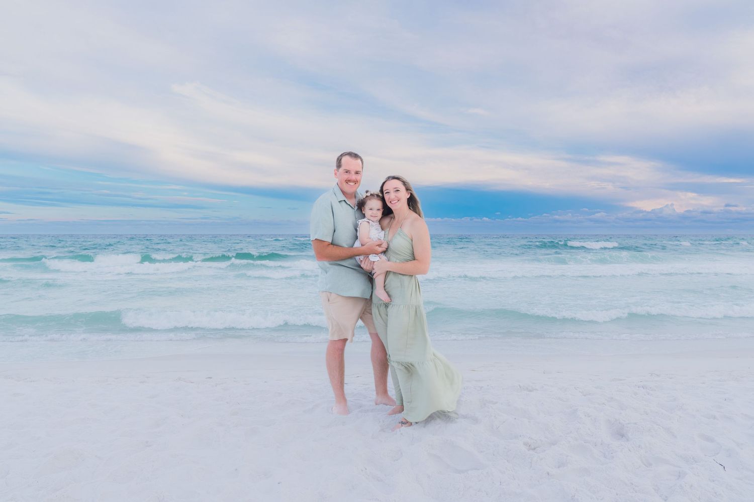 Family of three poses on a white sand beach at sunset, waves crashing behind them in soft pastel light.