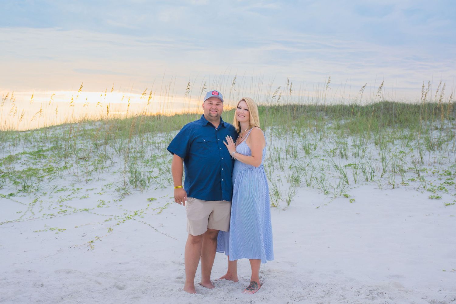 Couple posing together on white sand beach dunes at sunset, woman in blue dress and man in navy shirt.
