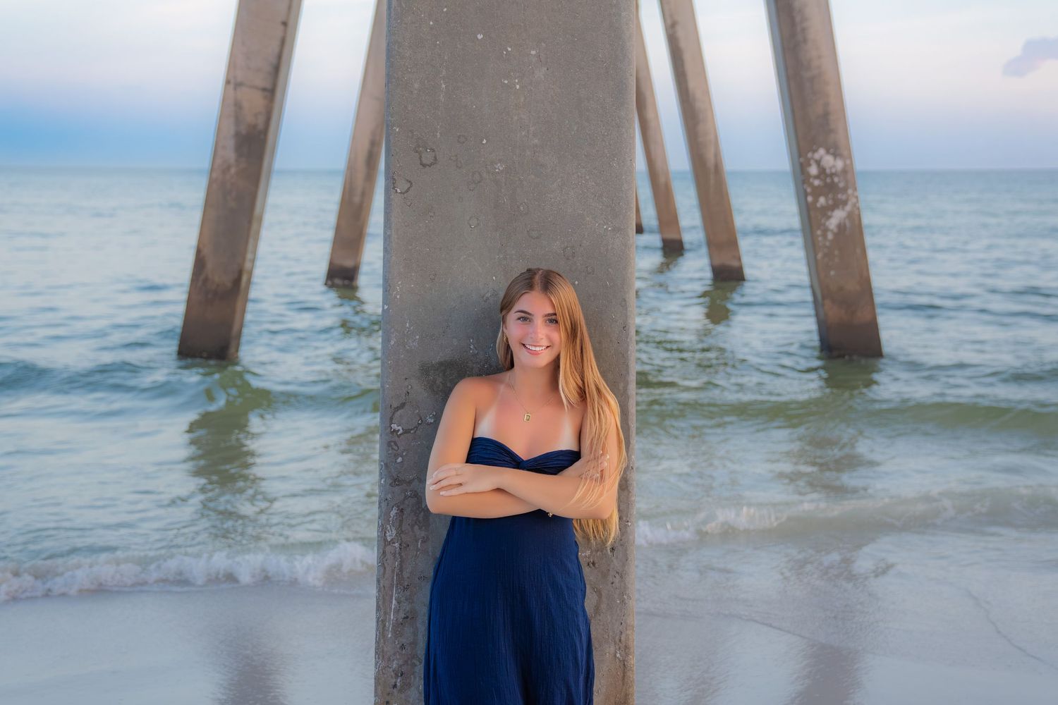 Young woman in navy dress leaning against pier pillar on sandy beach with ocean waves in background.