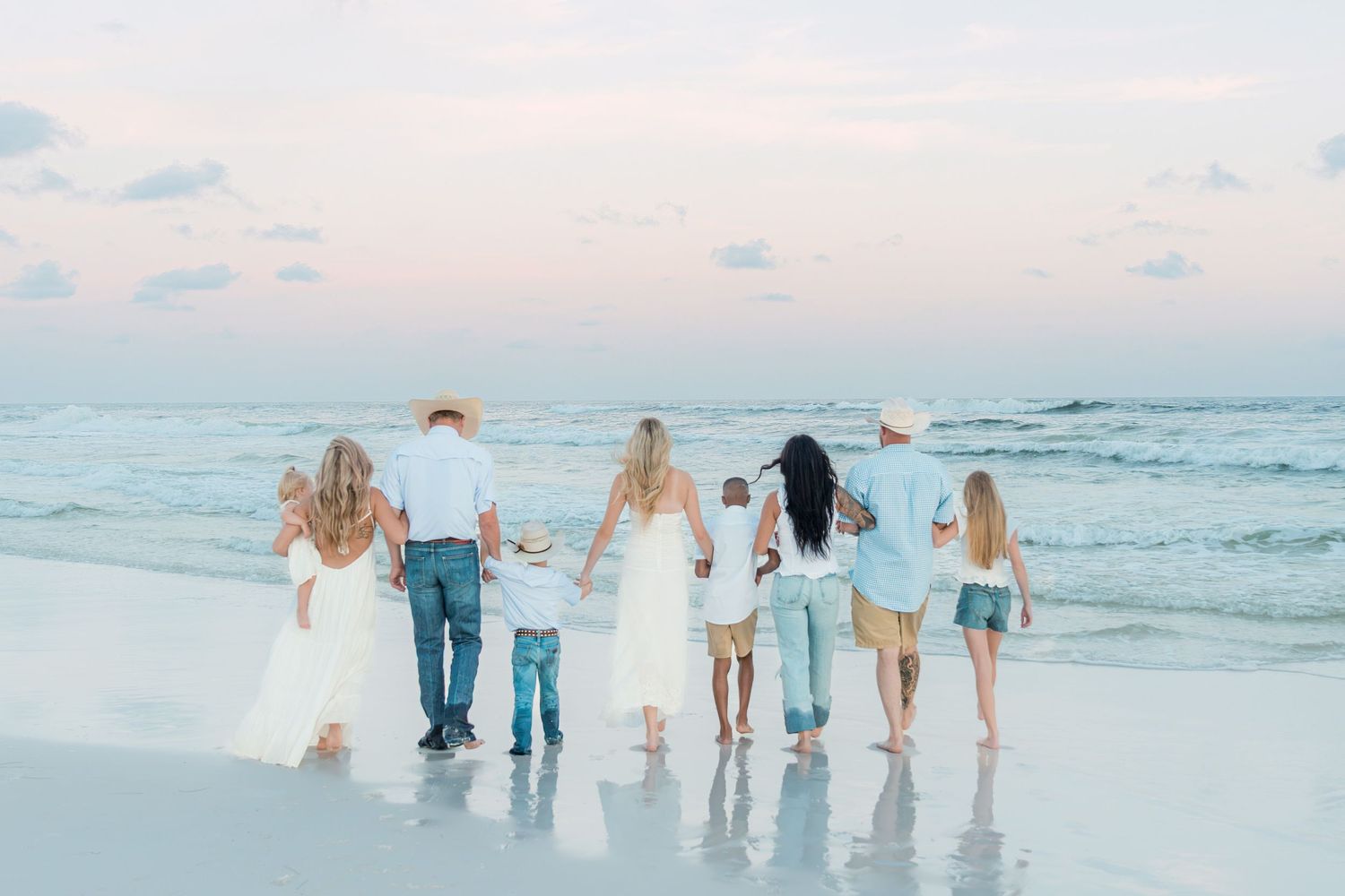 Large family holding hands walking along a sandy beach at sunset in matching white and blue outfits.