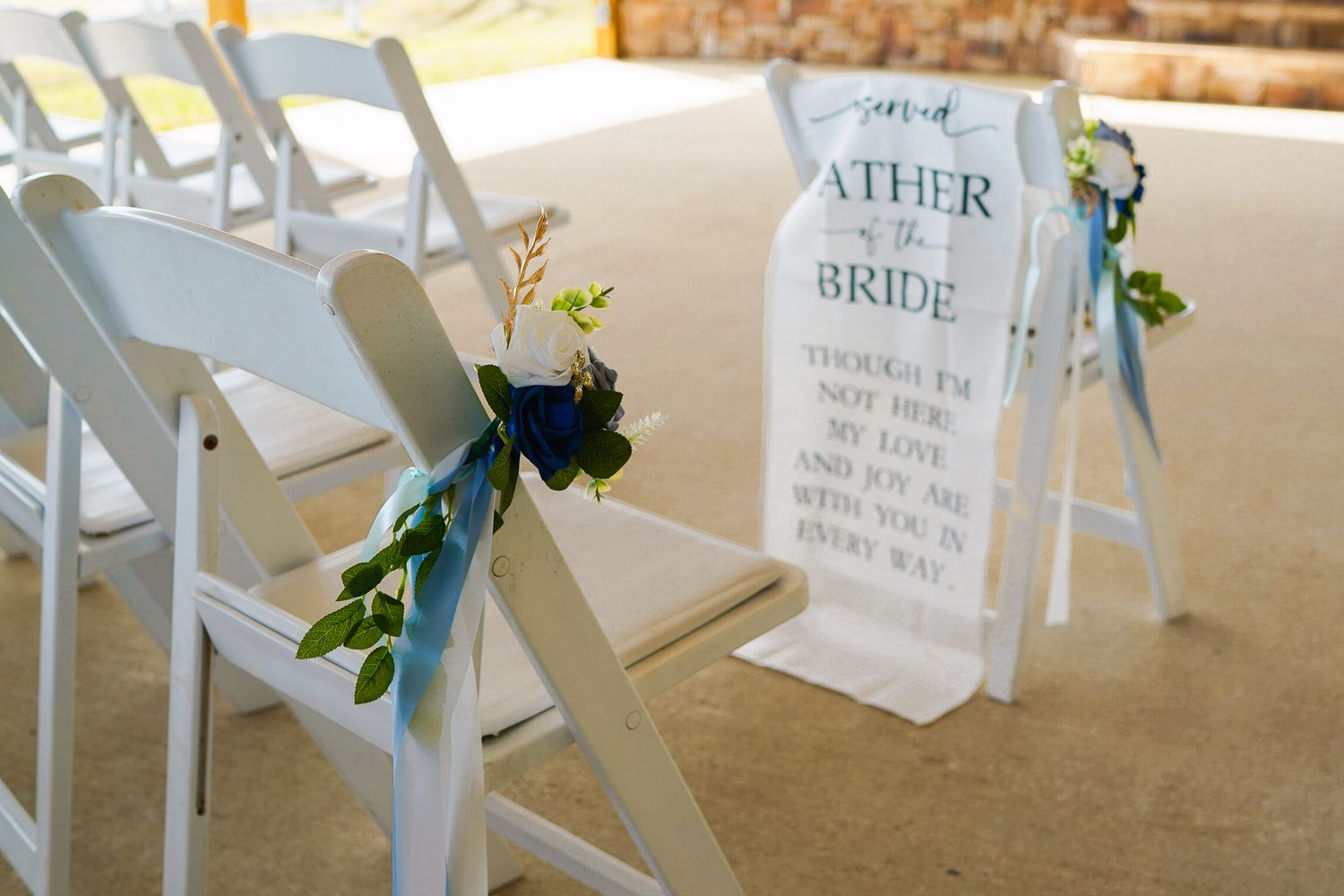 White wedding chairs with blue floral ribbons beside a Father of the Bride memorial sign at an outdoor ceremony.