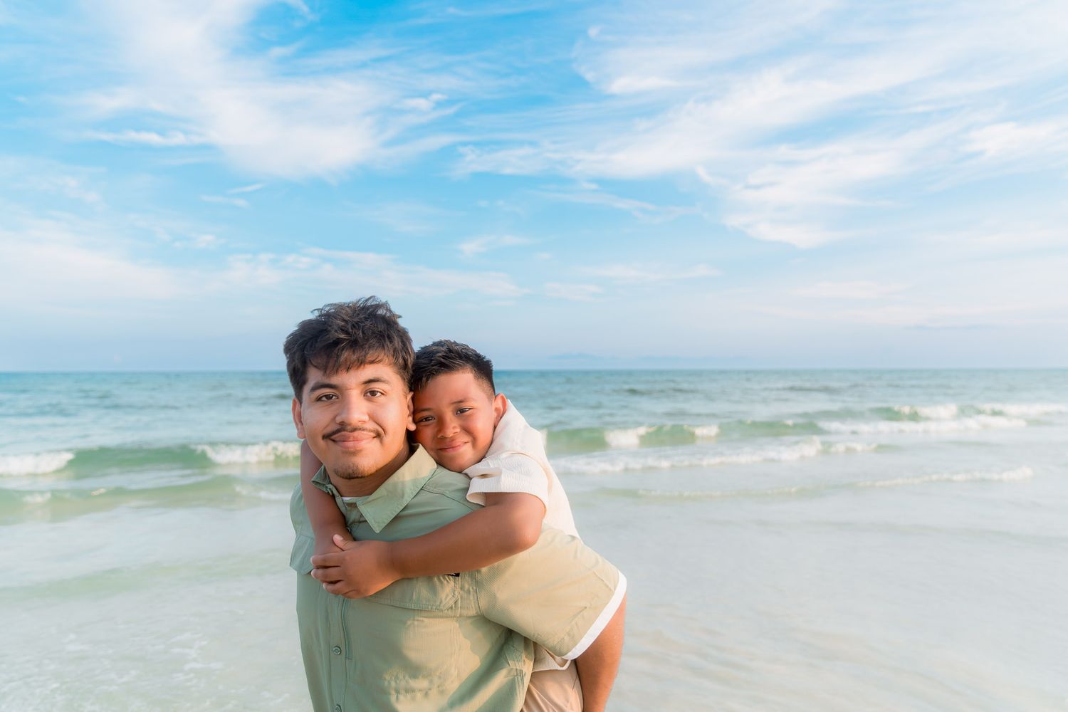 Man giving a child a piggyback ride on a sunny beach with blue skies and ocean waves in the background.