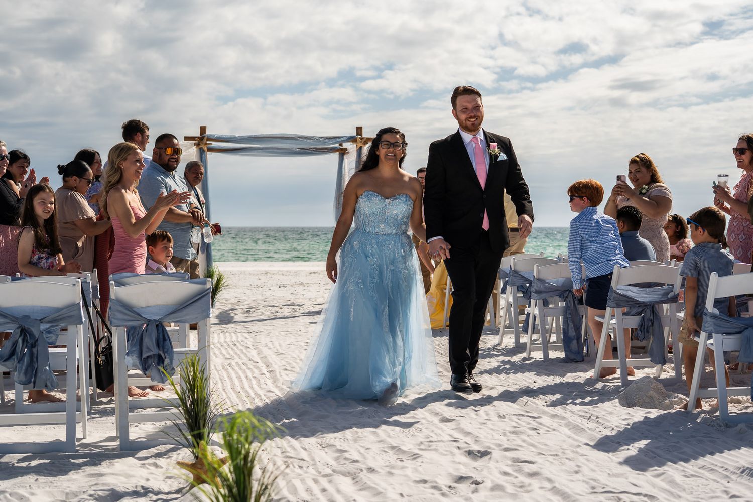 Newlyweds walk back down the aisle after beach wedding ceremony on white sandy shore.