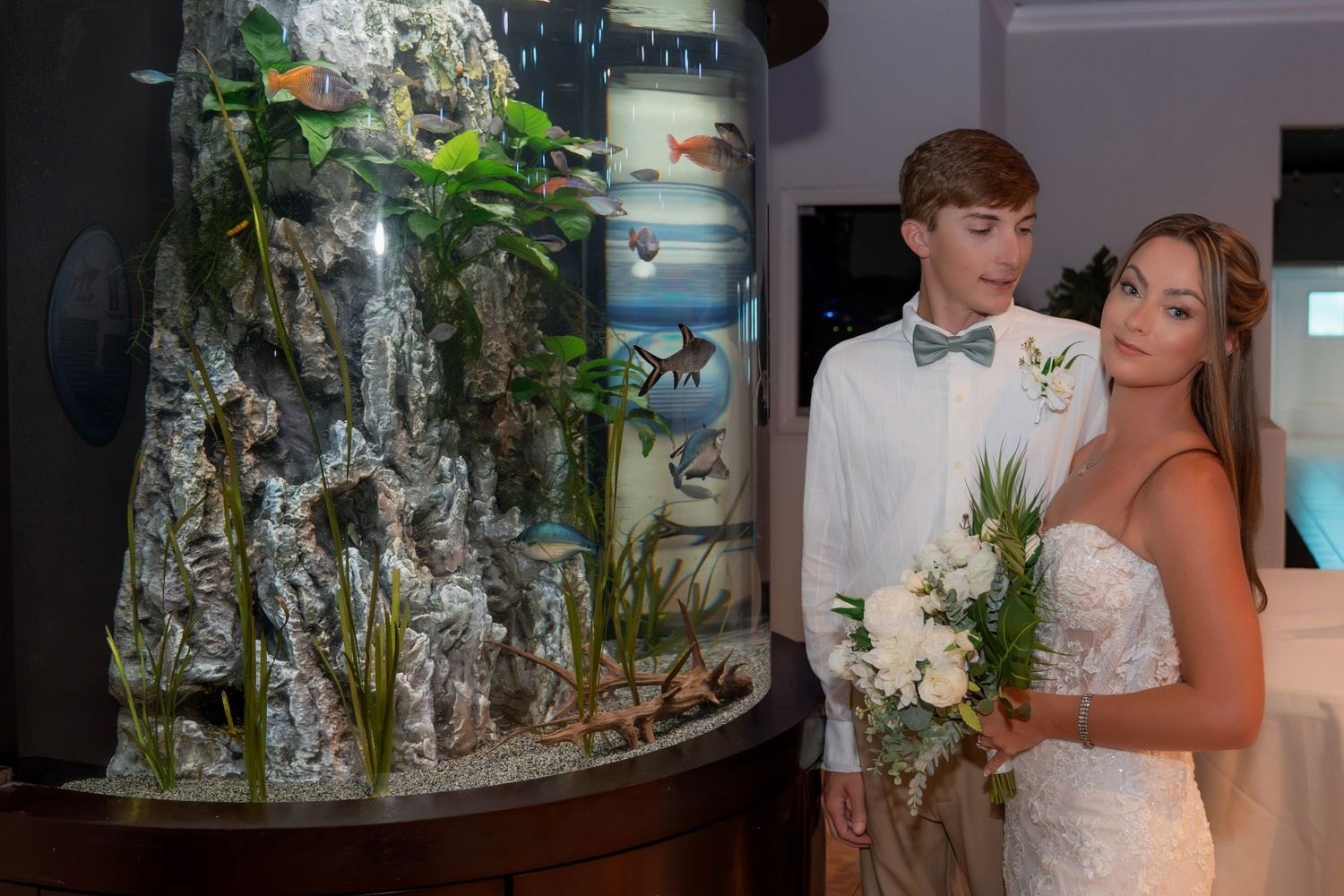 Newlywed couple in wedding attire admiring a large decorative aquarium with fish and rock formations.