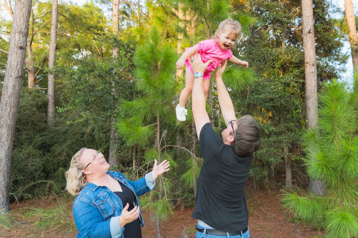 A joyful family plays outdoors as a man tosses a young girl in the air while a woman watches among tall pine trees.
