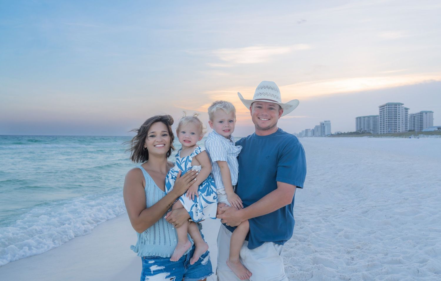 Family of four posing on a white sand beach at sunset with calm turquoise water and coastal buildings in the background.
