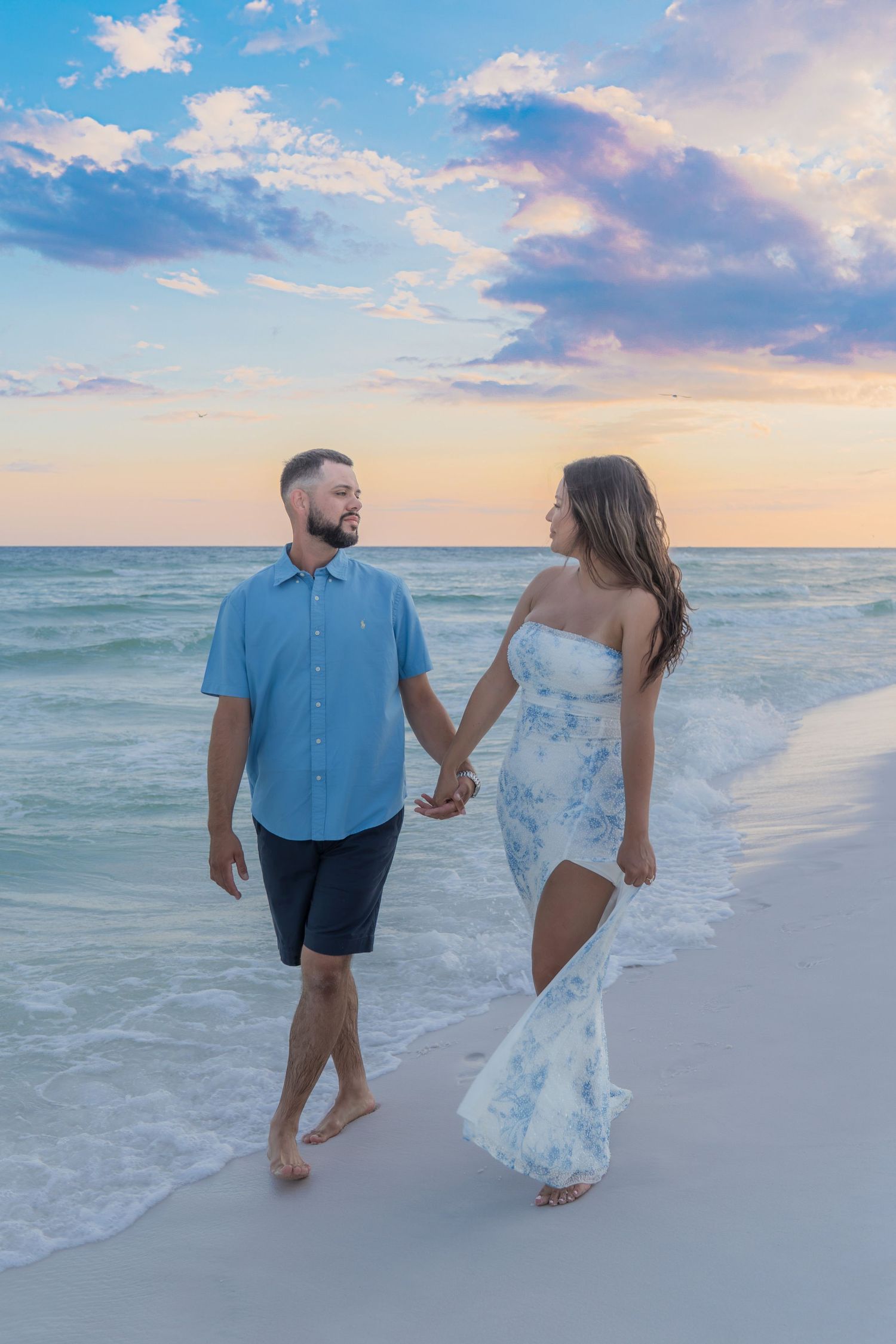Couple holds hands walking along shoreline at sunset, woman in floral dress, man in blue shirt and dark shorts.