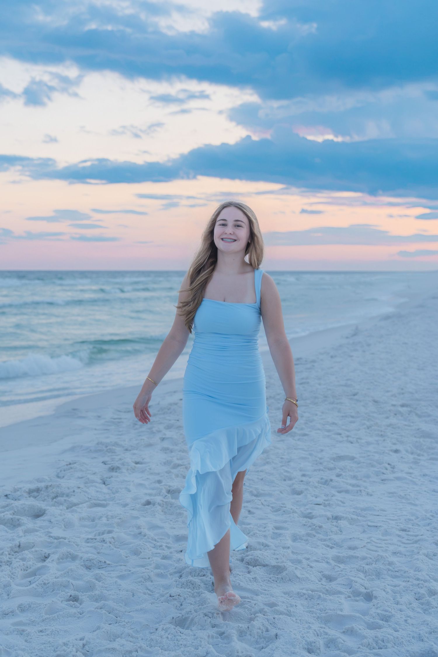 Young blonde woman in light blue ruffle dress walking barefoot on white sand beach at sunset.