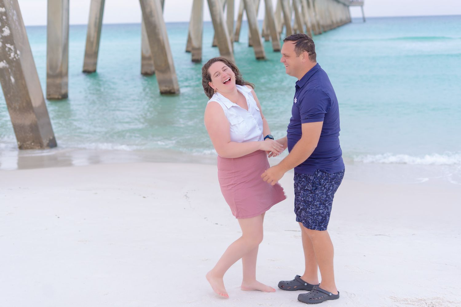 A laughing couple holds hands playfully on a white sand beach near a wooden pier at the shore.