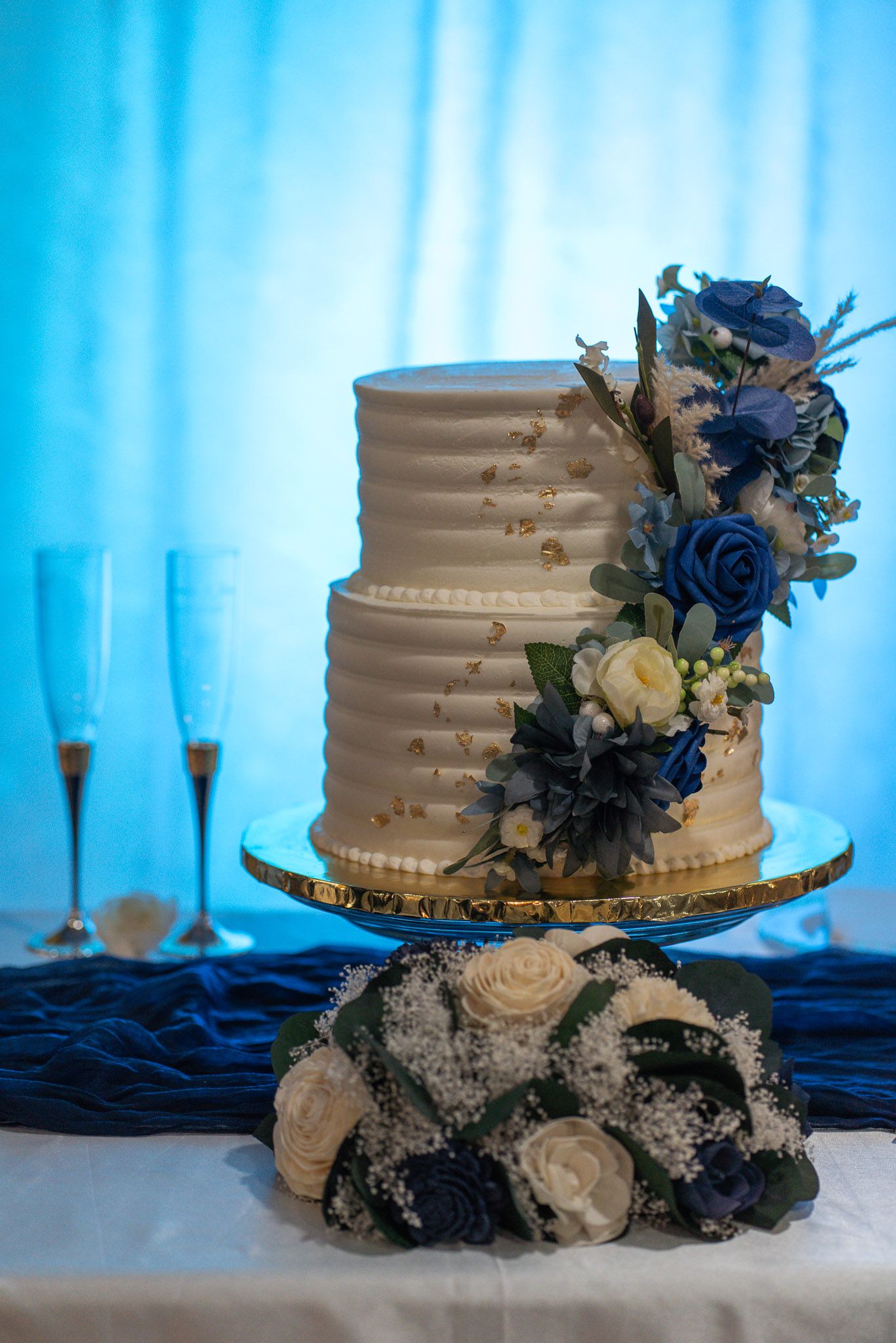 Elegant two-tier white wedding cake decorated with blue and white roses on a gold stand with champagne flutes.