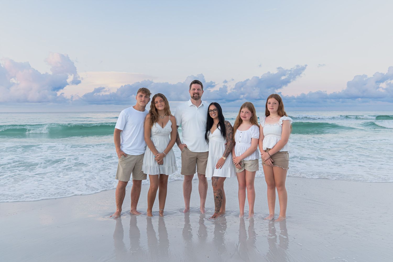 Family of six posing together on a sandy beach at sunset with turquoise waves and pastel sky behind them.