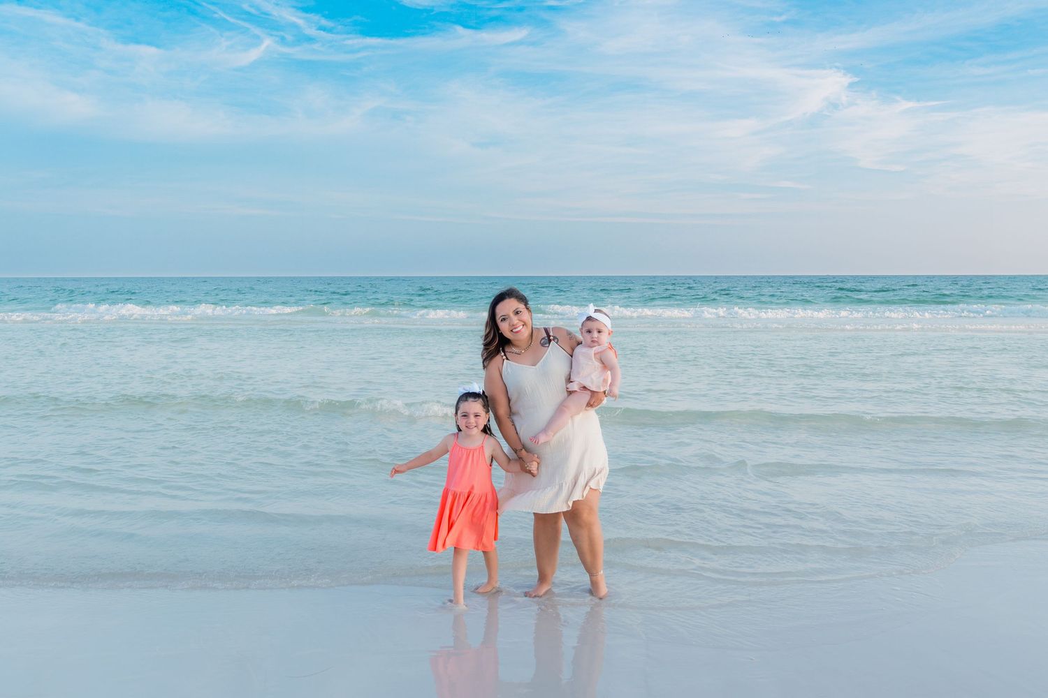 A mother and two young daughters pose happily on a sunny beach with turquoise ocean waves behind them.