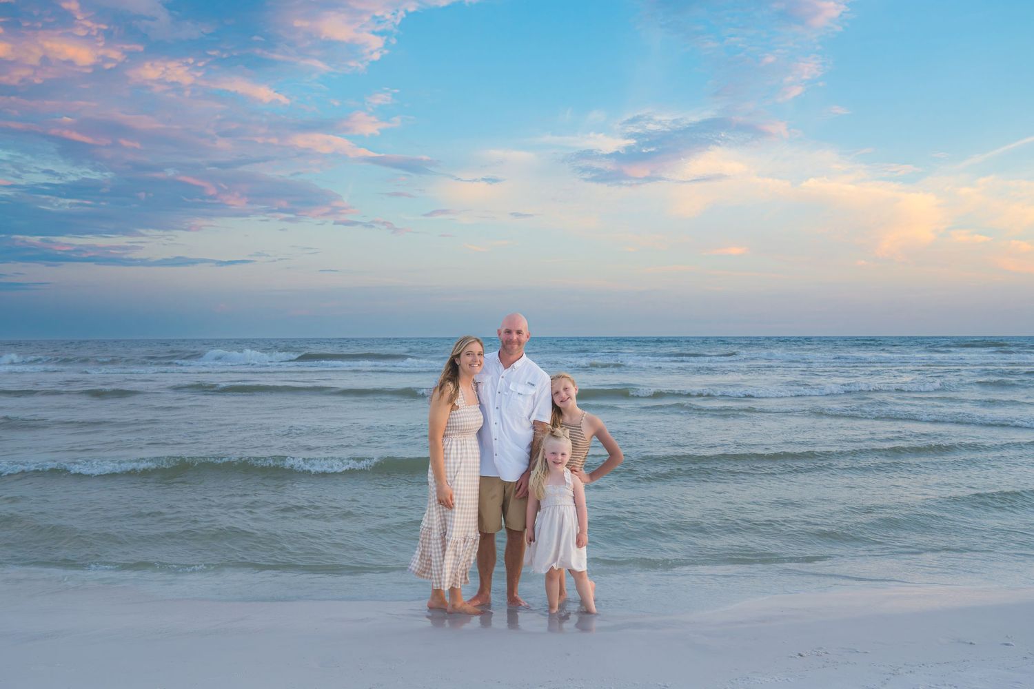 Family of four posing on a sandy beach at sunset with turquoise waves and colorful sky behind them.