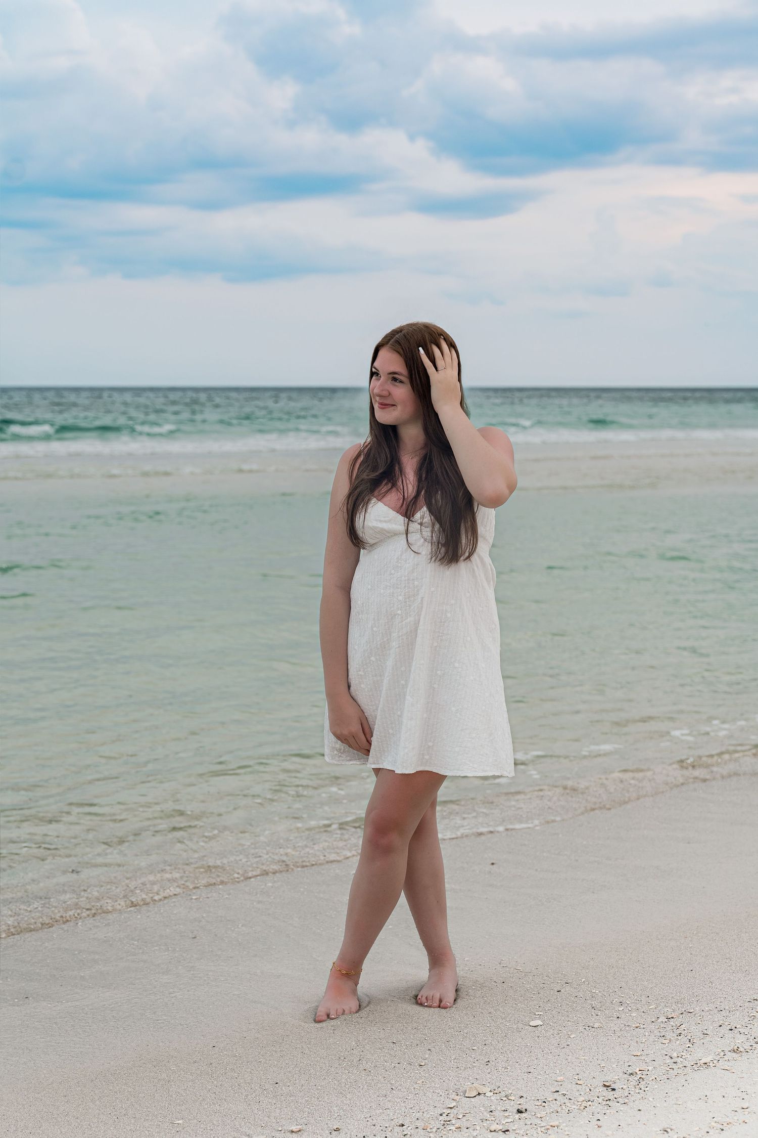 Young woman in white sundress walking barefoot on a white sand beach with turquoise ocean waves behind her.