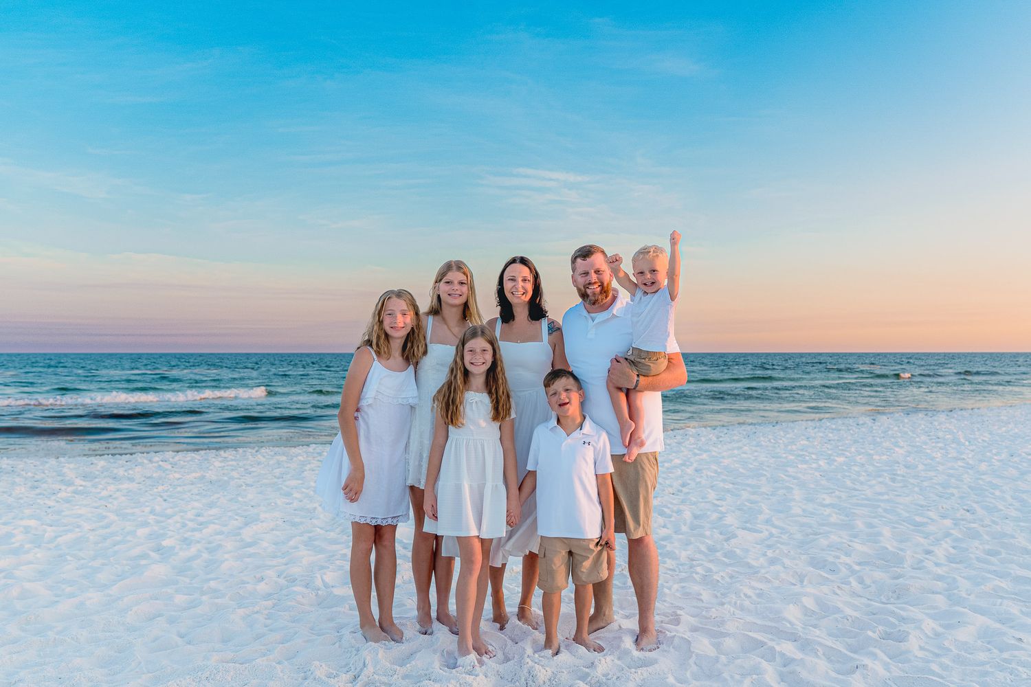Family of seven posing together on a sandy beach at sunset, wearing coordinated white and khaki outfits.