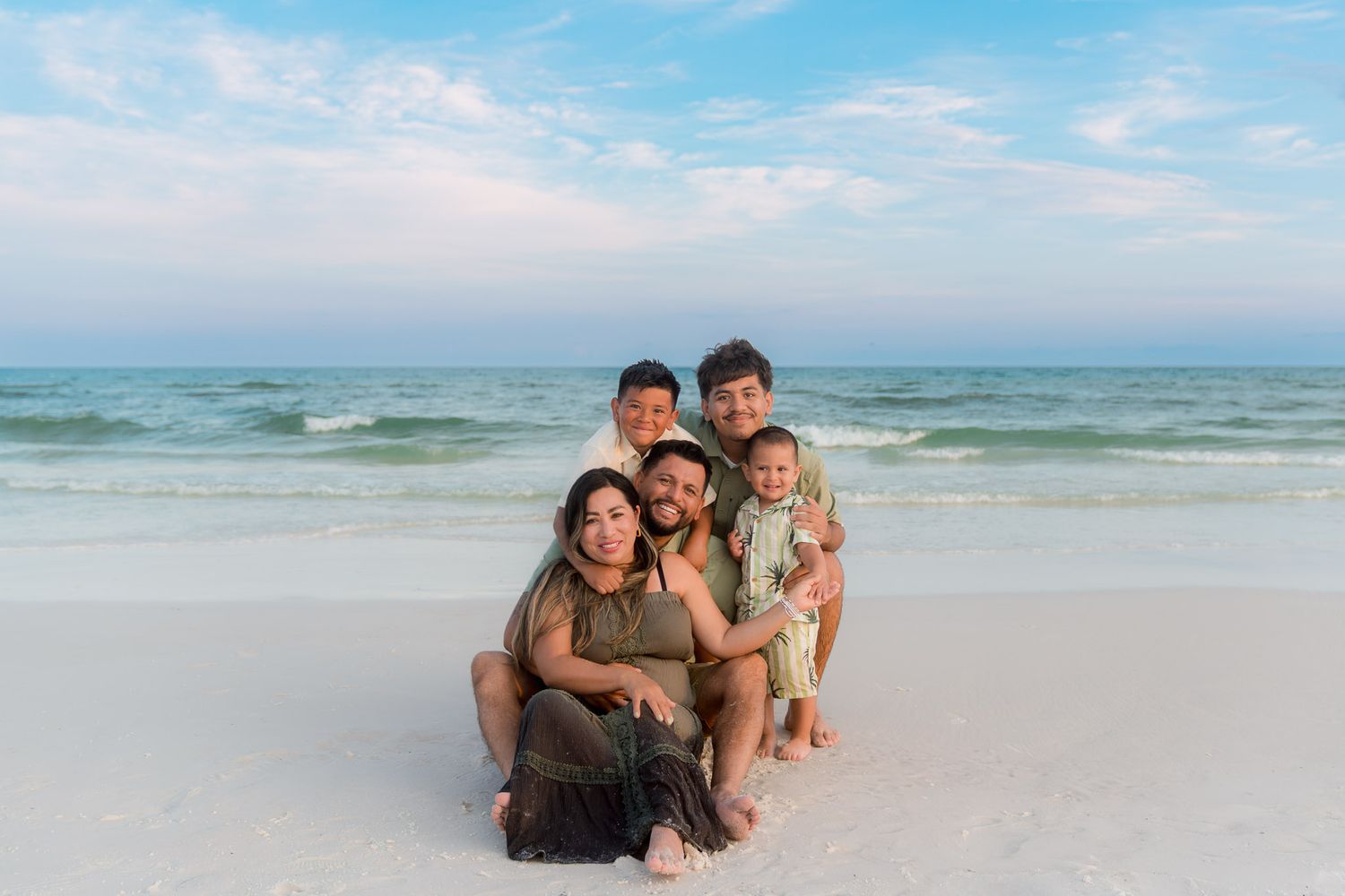 Happy family of five posing together on a white sandy beach with ocean waves behind them.