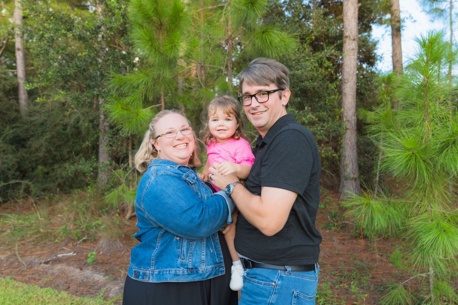 Happy family of three posing outdoors among pine trees, parents holding their toddler daughter.