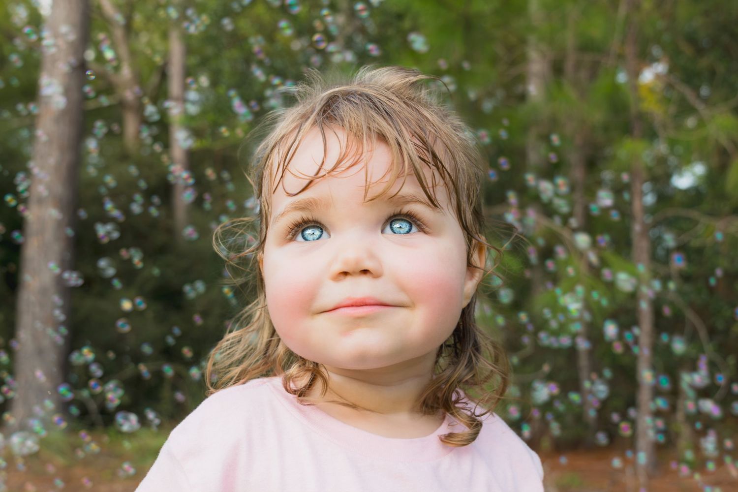 Cute toddler girl with blue eyes looks up at bubbles floating around her in an outdoor setting.