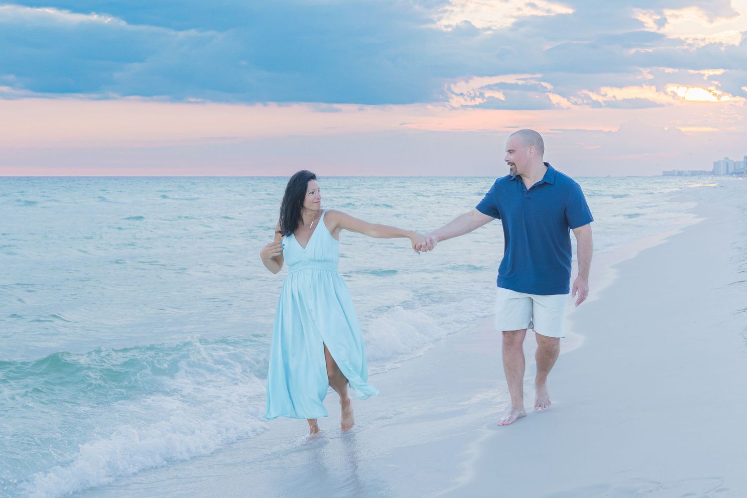 Couple holding hands walking along a sandy beach at sunset, woman in light blue dress, man in navy shirt and white shorts.