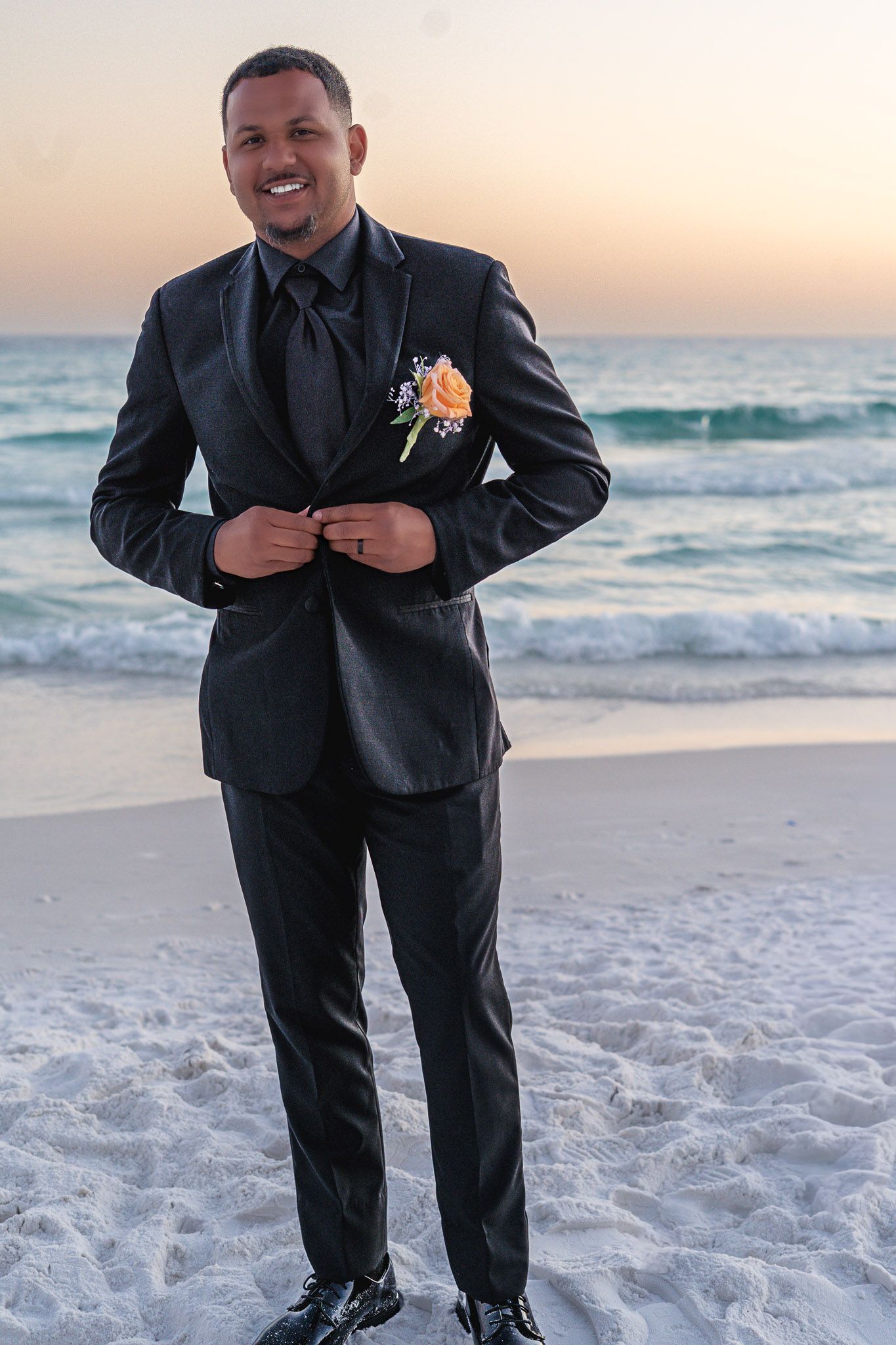 Smiling groom in black tuxedo with orange boutonniere stands on sandy beach at sunset with ocean waves behind him.