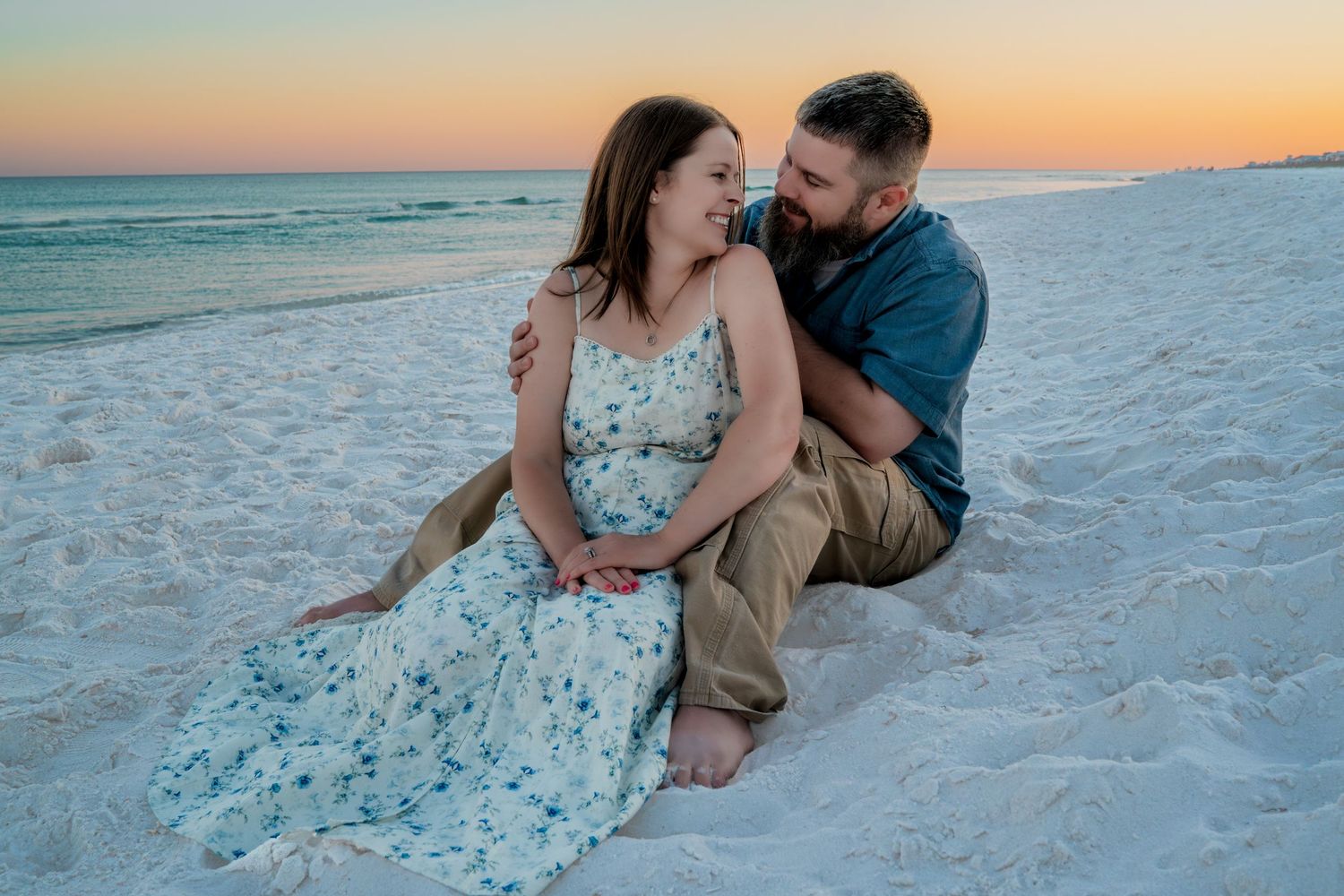 Couple sitting on white sand beach at sunset, woman in floral dress, man in blue shirt, sharing a romantic moment.