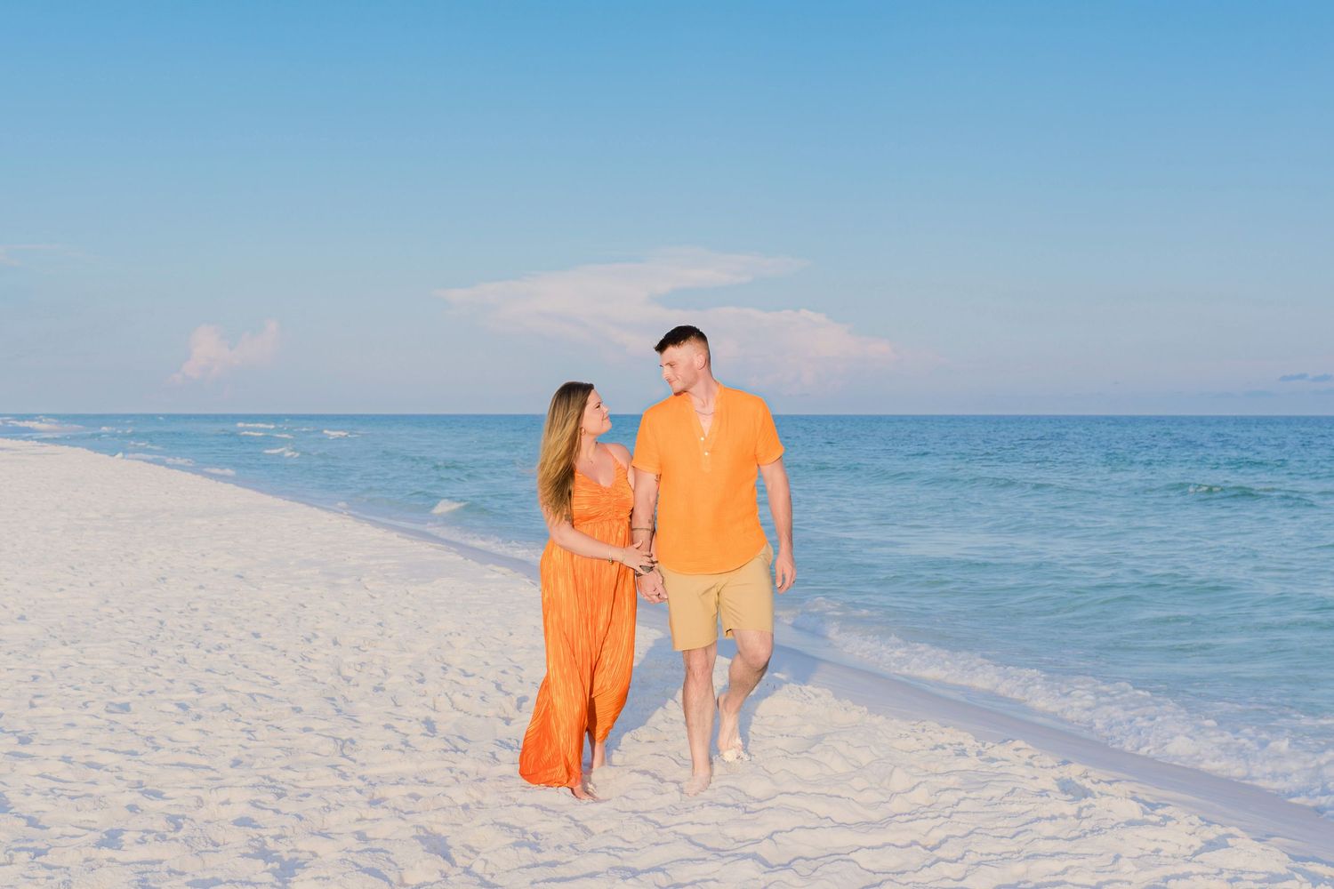 Couple in matching orange outfits walking hand-in-hand on a white sand beach with turquoise water.