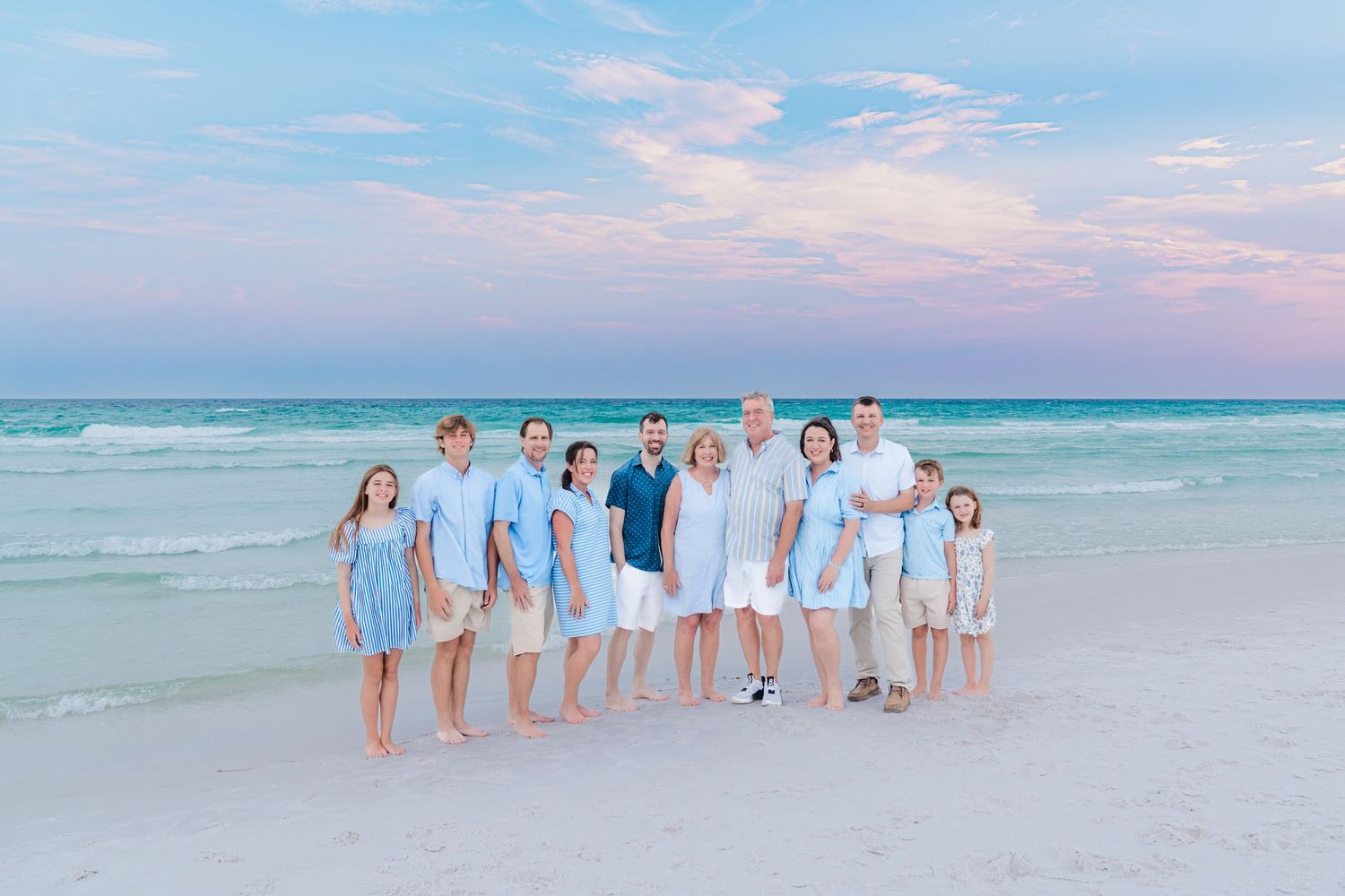 Large family posing together on a sandy beach at sunset with turquoise water and pink sky backdrop.