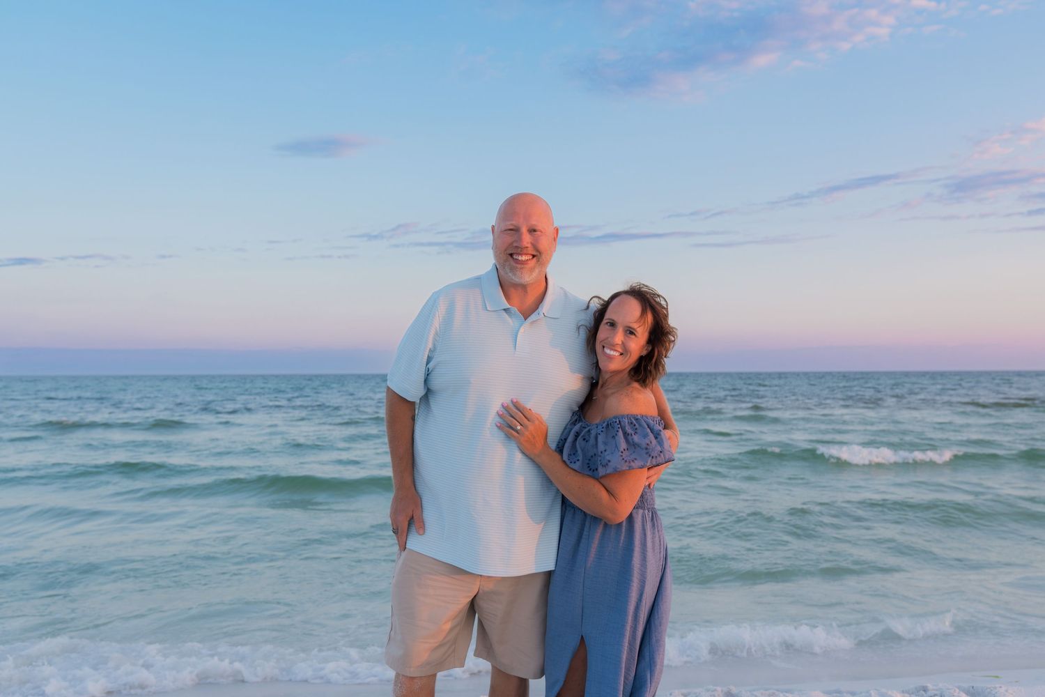 Couple smiling together on a sandy beach at sunset with calm turquoise ocean waves in the background.