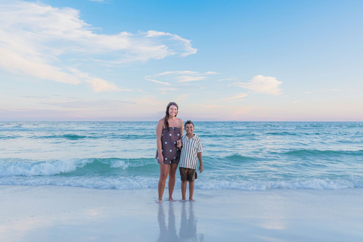 Two people standing together on a sandy beach with turquoise ocean waves and pastel sky behind them.