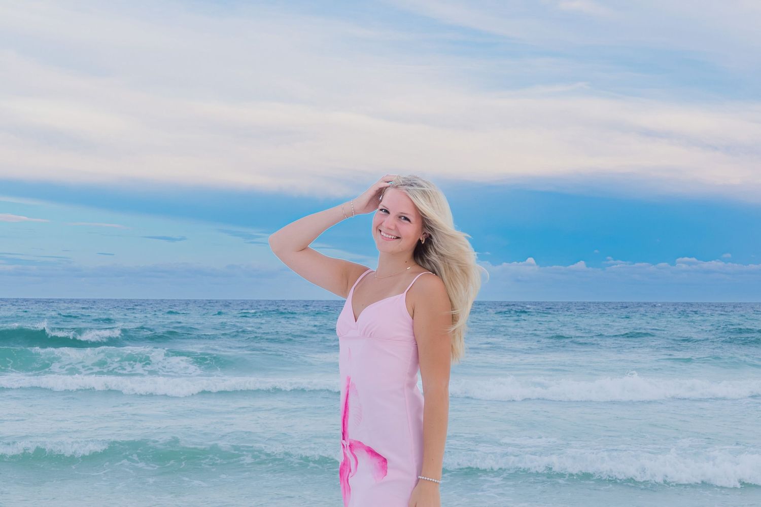 Smiling blonde woman in pink dress standing on a tropical beach with turquoise ocean waves and cloudy sky.