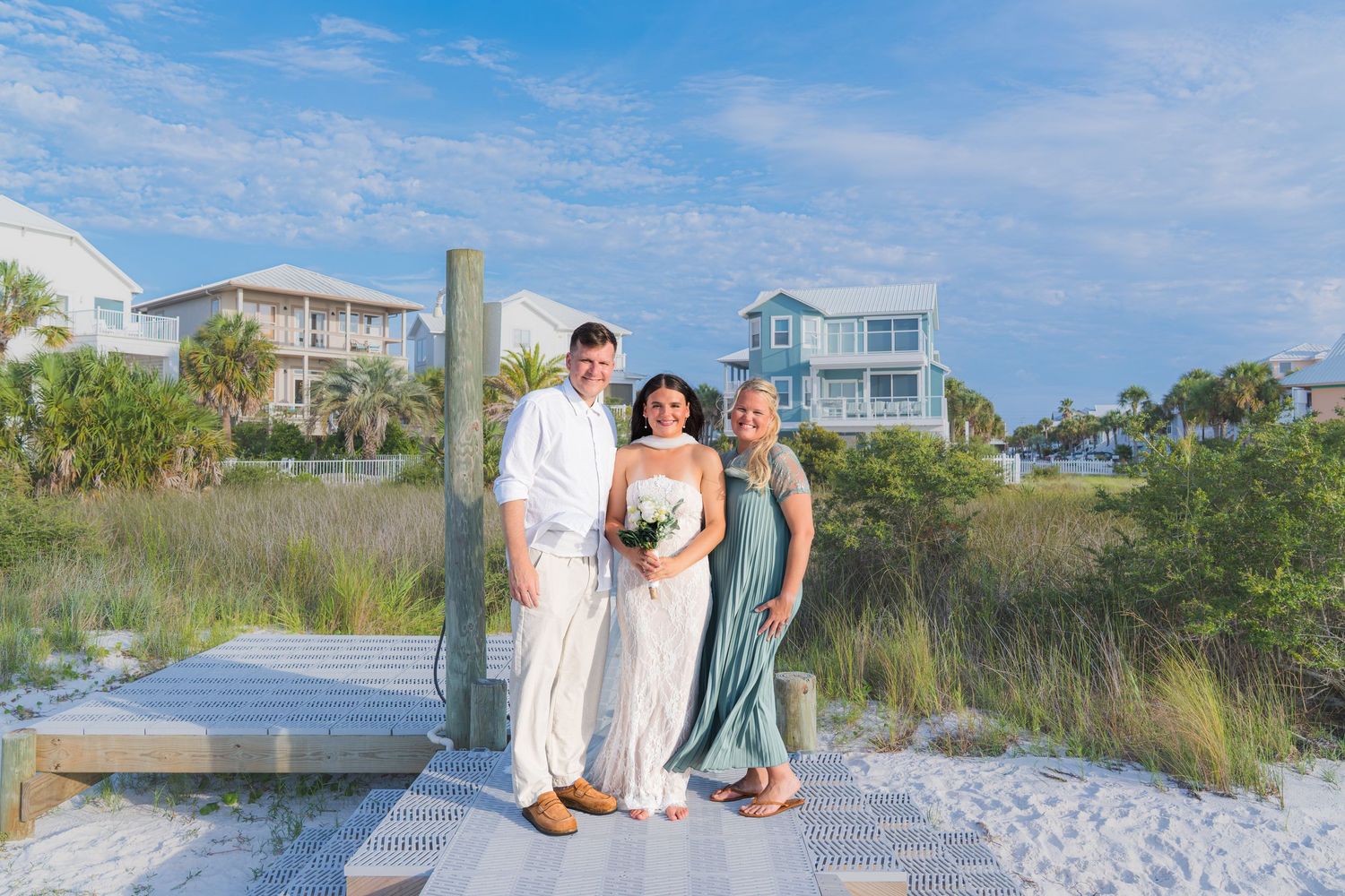 Bride and groom pose with female guest on sunny beach boardwalk near coastal homes.