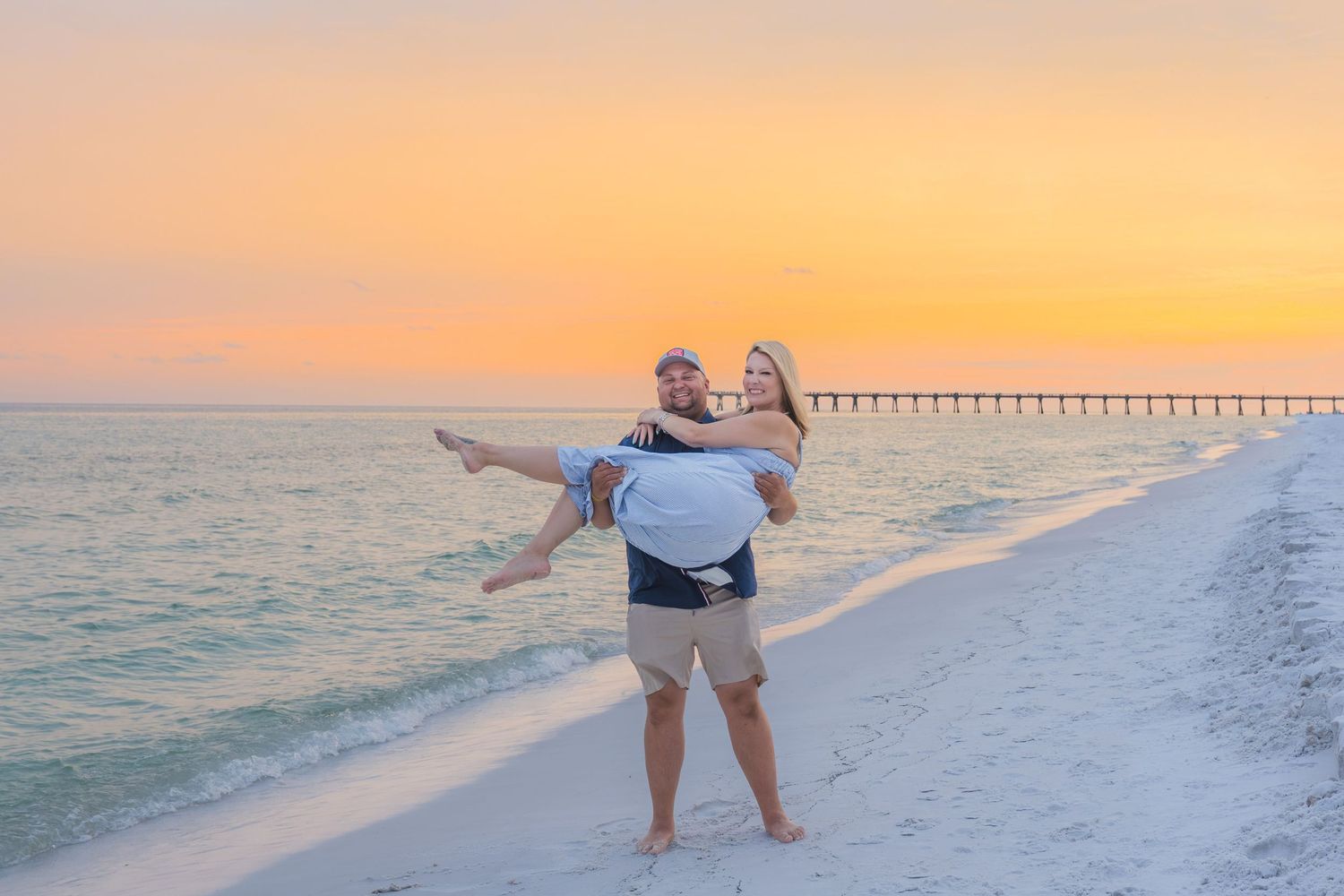 Man carrying woman on beach at golden sunset with pier visible in background.