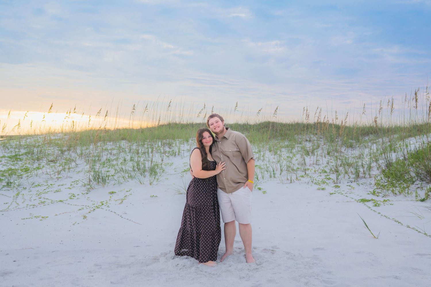 Couple embracing on white sand beach dunes at sunset with sea oats in background.