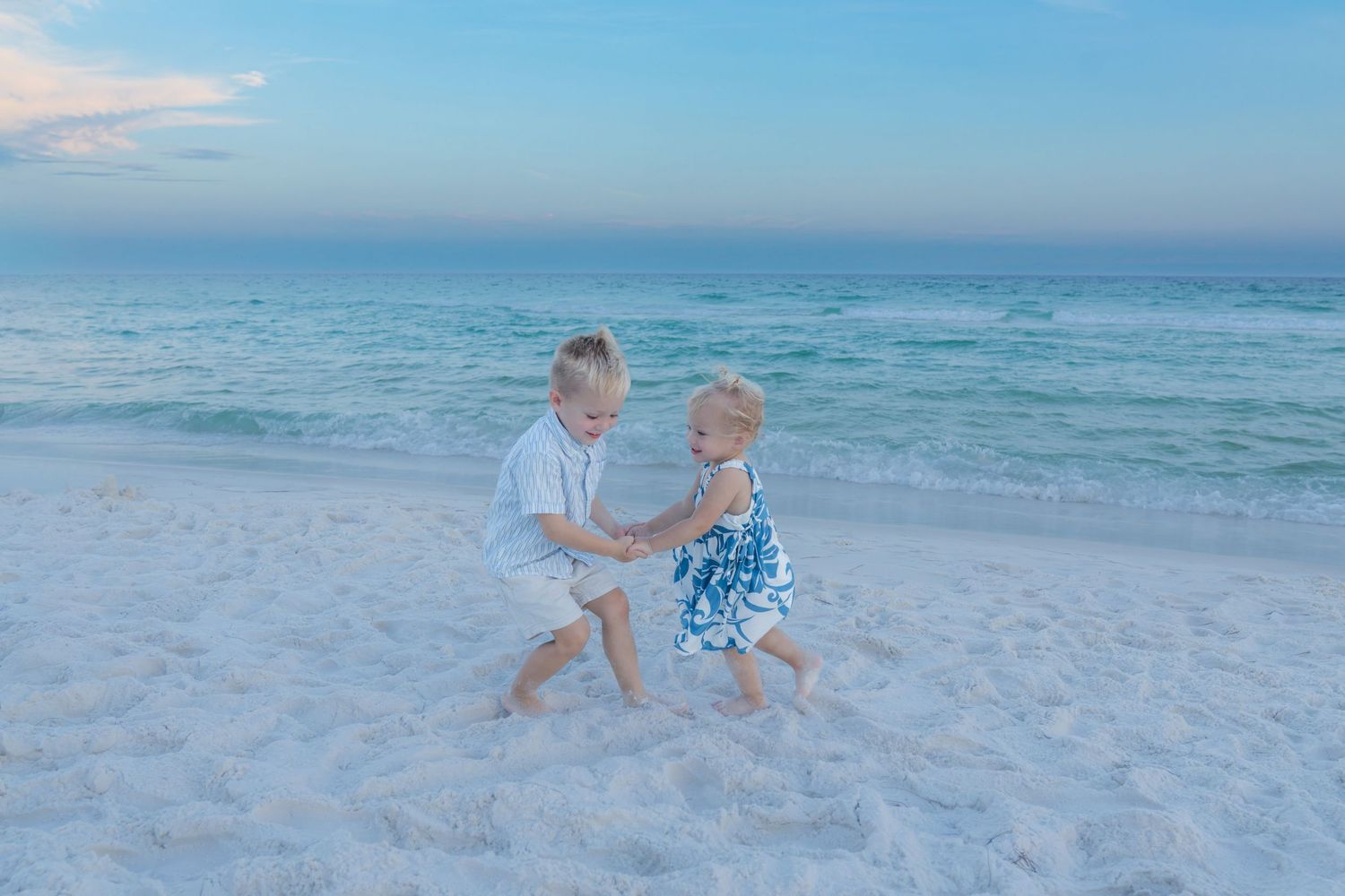 Two young children playing and dancing together on a white sandy beach with turquoise ocean waves at sunset.