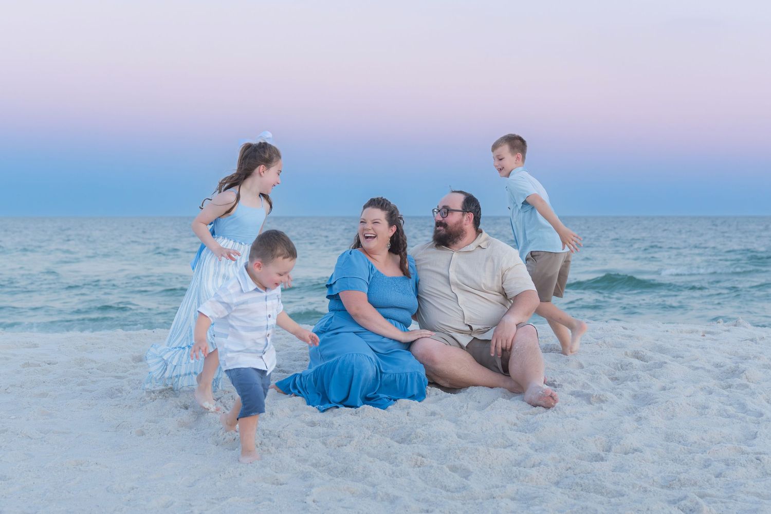 Family of five enjoying a beach sunset, parents sitting on white sand while children play near gentle ocean waves.
