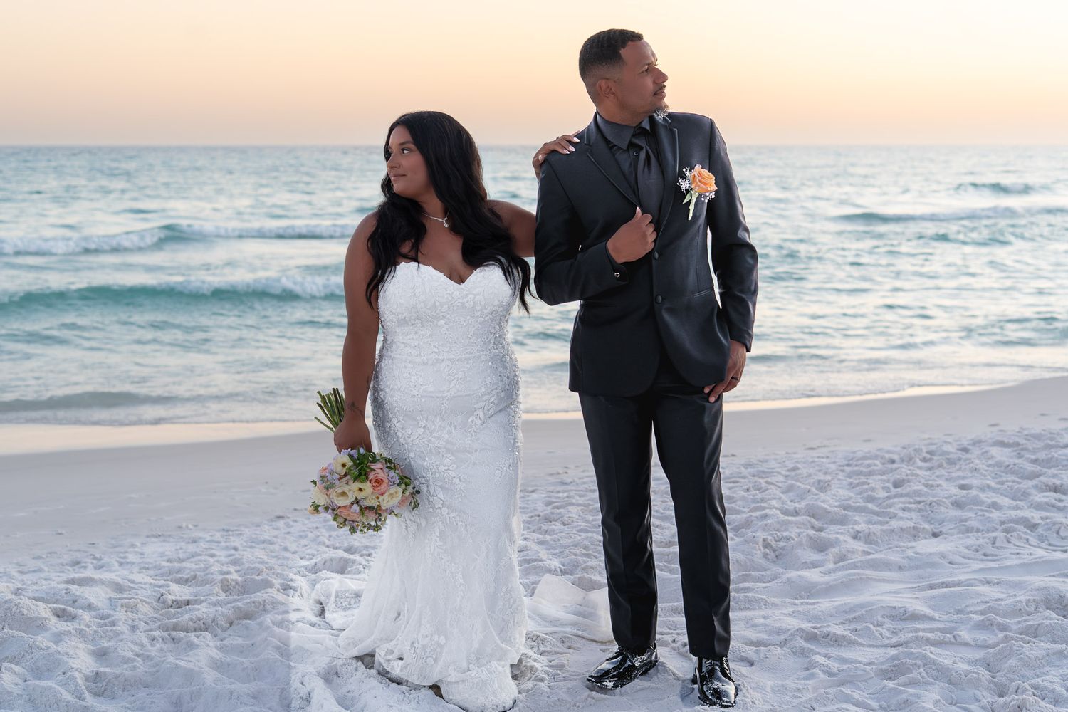 Newlywed couple posing on a sandy beach at sunset, bride in white gown holding bouquet, groom in dark suit.