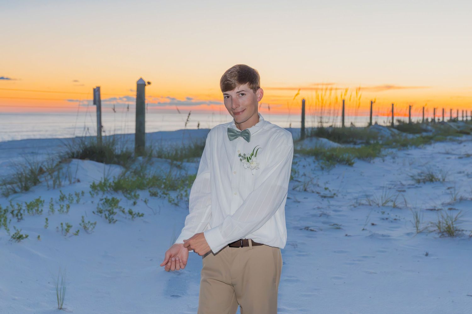 A young groom in a white shirt and khakis poses on a sandy beach at sunset with ocean views.