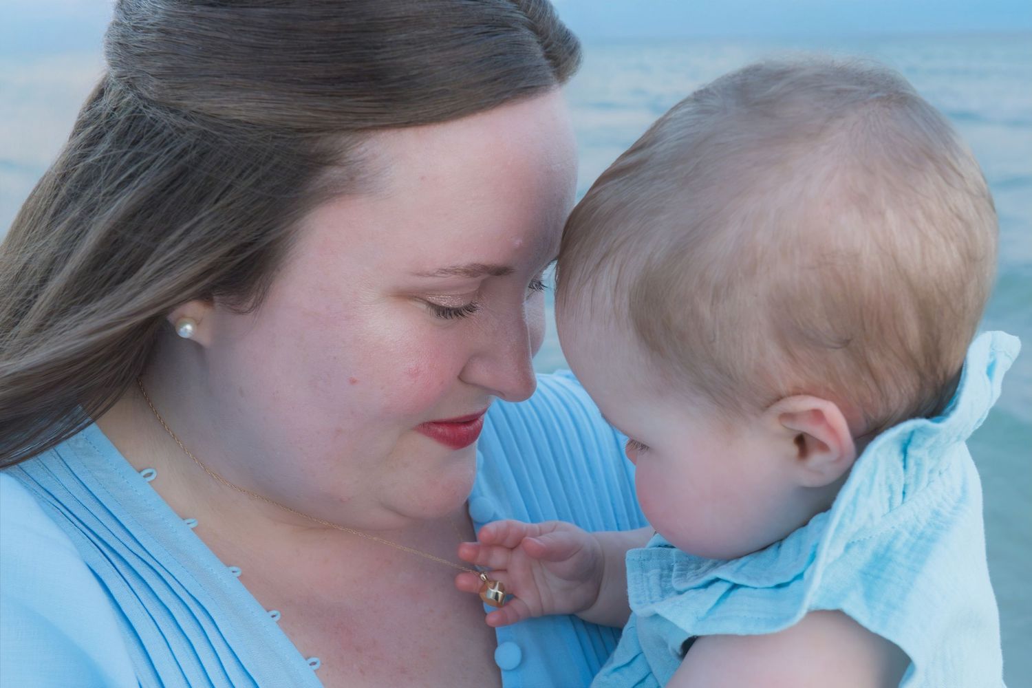 A mother in a blue dress tenderly touches foreheads with her baby at the beach.